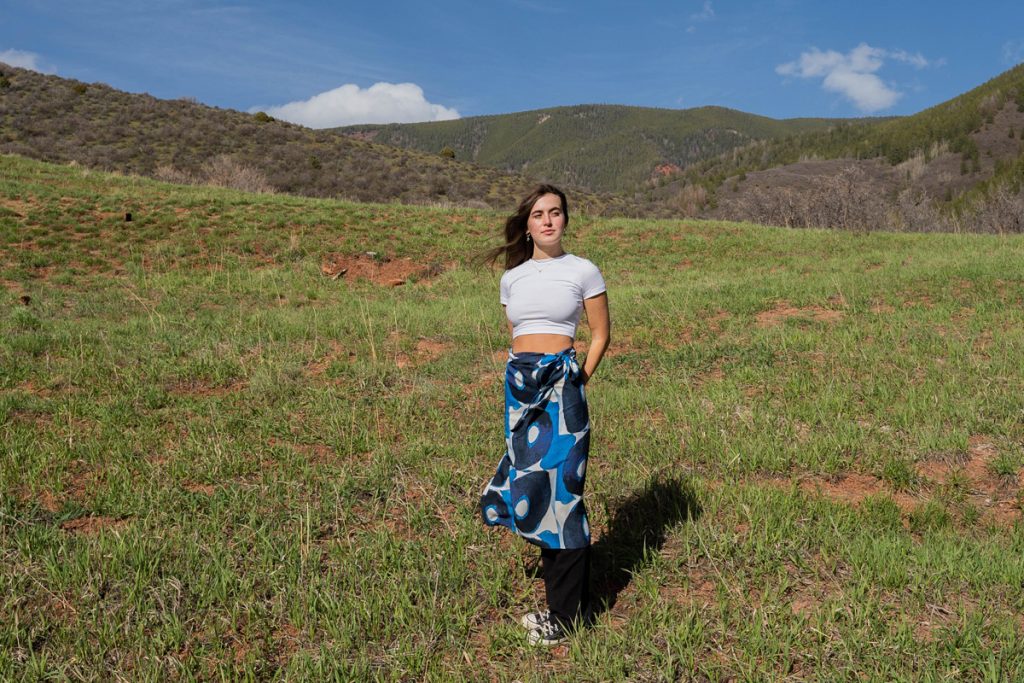 a young woman standing in a mountain meadow with a blue and white graphic patterned wrap around skirt and a white t shirt with hair blowing in the wind