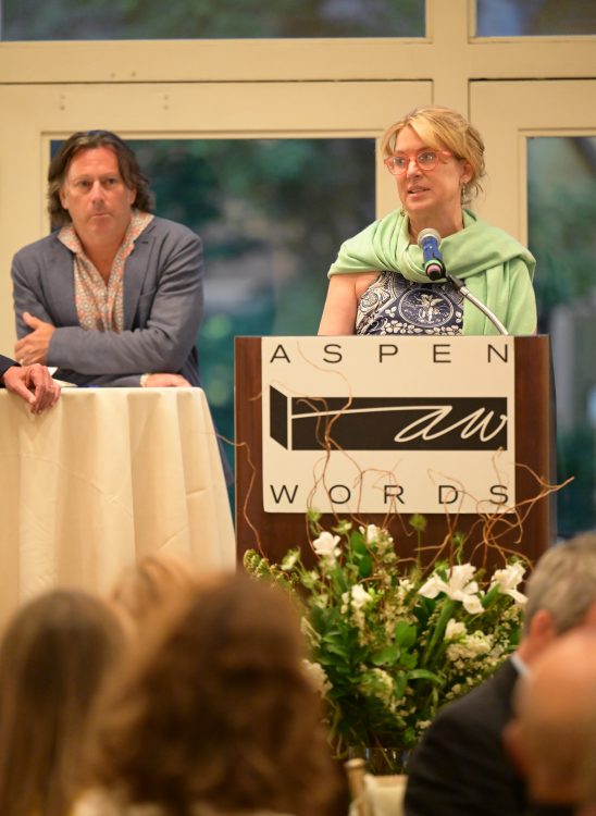 Isa Catto speaking on stage behind podium at Aspen Words event with husband, Daniel Shaw in background.